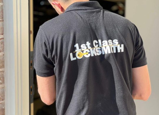 1st Class Locksmith technician working on a residential door lock, wearing a black uniform with yellow company branding in Richardson, TX